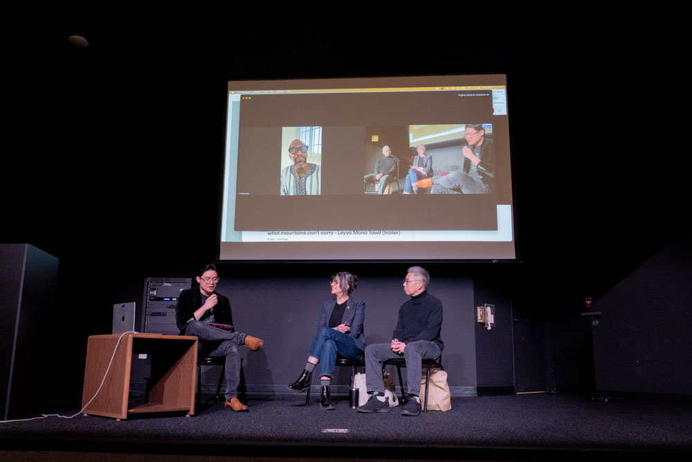Two panelist, the moderator on stage, with the final panelist on Zoom behind them.